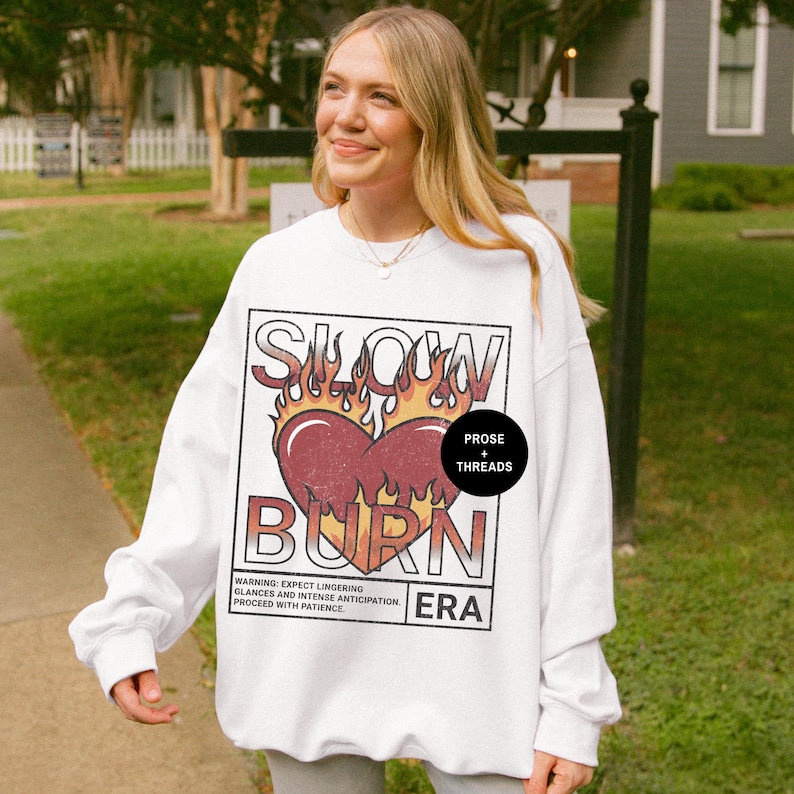 May include: White sweatshirt with a graphic print of a heart on fire with the text "Slow Burn" and "Prose + Threads" in a black circle. The text "ERA" is below the graphic.