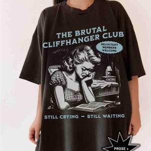 May include: Black t-shirt with a graphic featuring the text "THE BRUTAL CLIFFHANGER CLUB" and an illustration of a crying woman. The shirt also includes the phrases "RELUCTANT MEMBERS WELCOME" and "STILL CRYING - STILL WAITING".