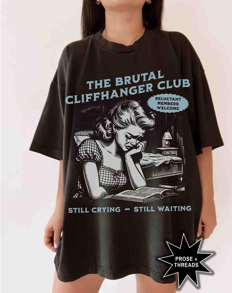 May include: Black t-shirt with a graphic featuring the text "THE BRUTAL CLIFFHANGER CLUB" and an illustration of a crying woman. The shirt also includes the phrases "RELUCTANT MEMBERS WELCOME" and "STILL CRYING - STILL WAITING".