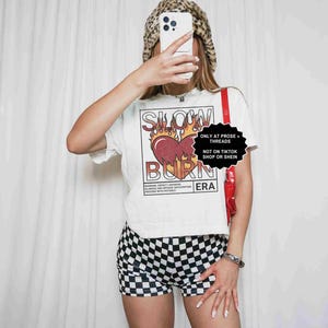 May include: A white t-shirt with a graphic that says "SLOW BURN ERA" with a heart and flames design. The person is wearing black and white checkered shorts and a leopard print hat. A red strap is visible.