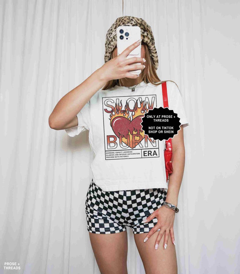 May include: A white t-shirt with a graphic that says "SLOW BURN ERA" with a heart and flames design. The person is wearing black and white checkered shorts and a leopard print hat. A red strap is visible.