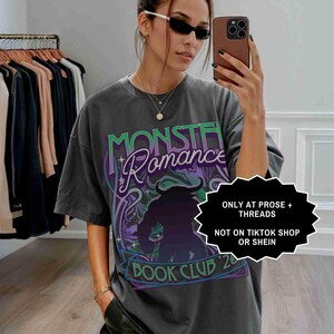 Op de afbeelding: Donkergrijs t-shirt met de tekst "MONSTER ROMANCE BOOK CLUB '20" in groen en paars. Het shirt heeft een afbeelding van een gehoornde figuur. De tekst "ONLY AT PROSE + THREADS NOT ON TIKTOK SHOP OR SHEIN" is ook zichtbaar.