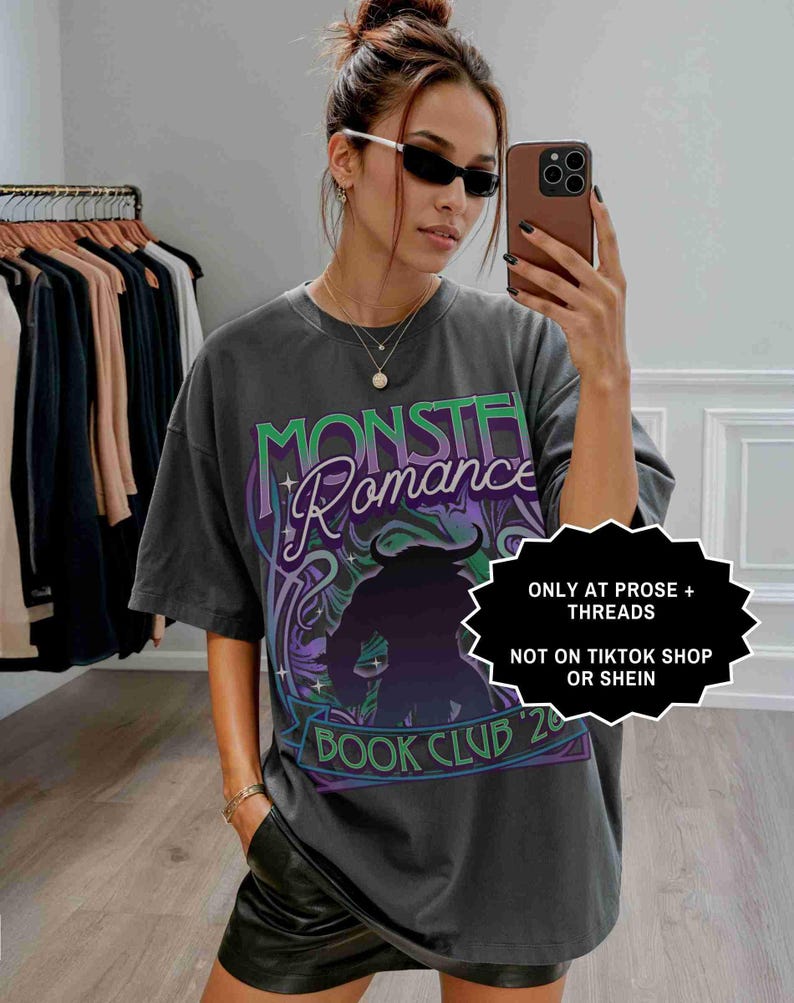 Op de afbeelding: Donkergrijs t-shirt met de tekst "MONSTER ROMANCE BOOK CLUB '20" in groen en paars. Het shirt heeft een afbeelding van een gehoornde figuur. De tekst "ONLY AT PROSE + THREADS NOT ON TIKTOK SHOP OR SHEIN" is ook zichtbaar.