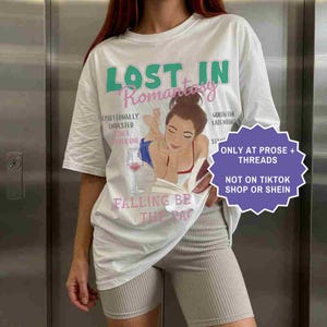 May include: White t-shirt with the text "LOST IN Romantasy" in green and pink. The shirt features an illustration of a woman reading with a glass of wine. Additional text includes "Emotionally Invested" and "Falling Be The Rac".