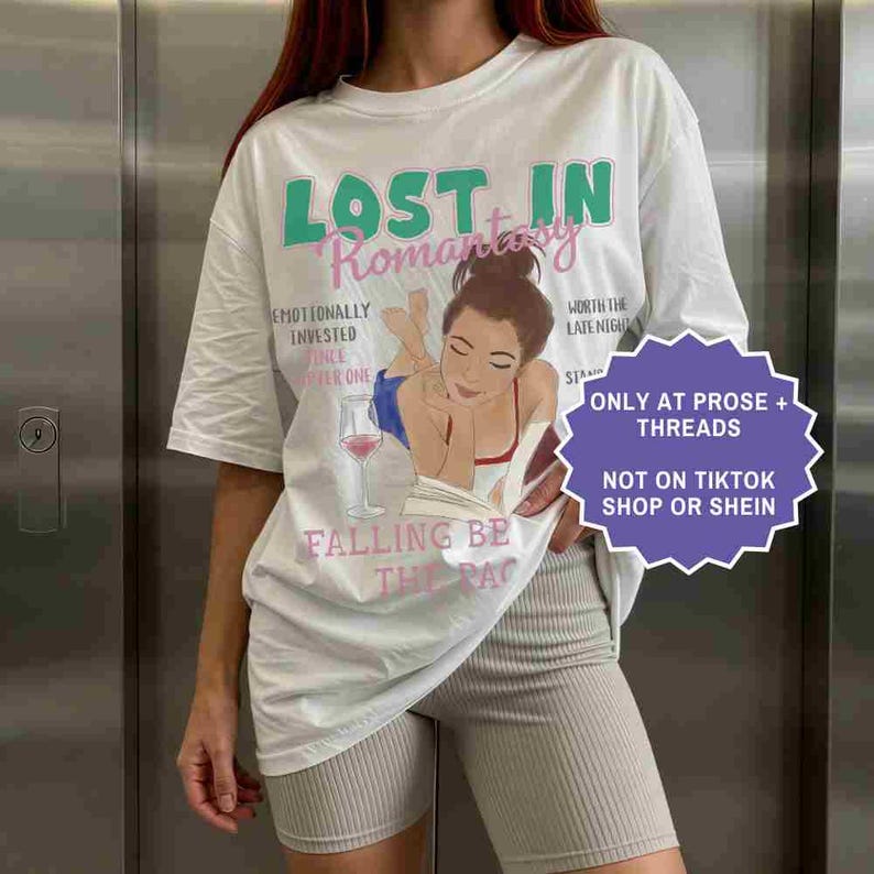 May include: White t-shirt with the text "LOST IN Romantasy" in green and pink. The shirt features an illustration of a woman reading with a glass of wine. Additional text includes "Emotionally Invested" and "Falling Be The Rac".