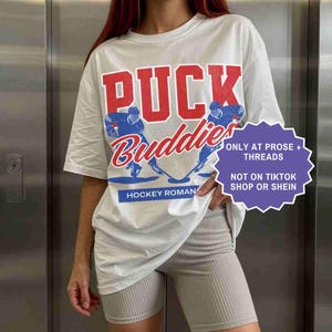 May include: White t-shirt with a hockey-themed graphic. The design features the words "PUCK Buddies" in red and blue, along with illustrations of hockey players. The text "HOCKEY ROMAN" is at the bottom.