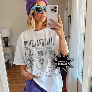 May include: White t-shirt with the text "ROMEO AND JULI" and illustrations of a sword, skull, angel wing, and rose. The shirt also has the word "Verona" printed on it. The person is wearing a purple beanie, blue sunglasses, and purple shorts.