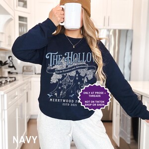 May include: Navy blue sweatshirt featuring a light blue graphic that reads "The Hollow Inn for Travelers and Adventurers." The design includes a tree with a house and the text "Merrywood Forest ESTD 2021." The sweatshirt is paired with grey sweatpants.