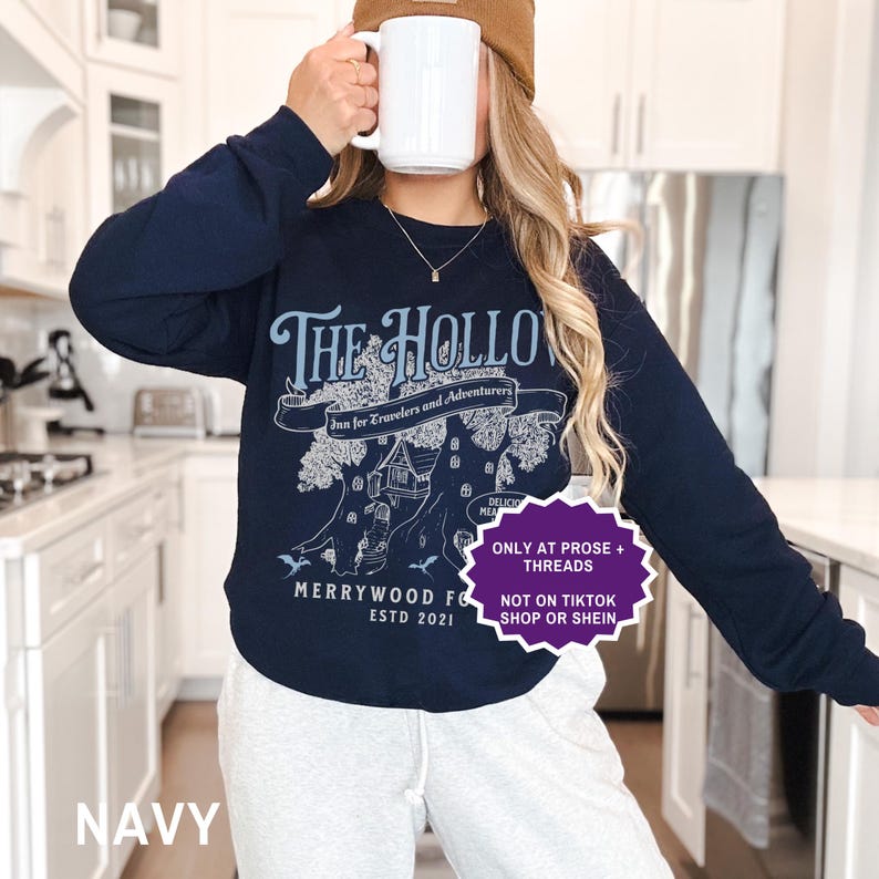 May include: Navy blue sweatshirt featuring a light blue graphic that reads "The Hollow Inn for Travelers and Adventurers." The design includes a tree with a house and the text "Merrywood Forest ESTD 2021." The sweatshirt is paired with grey sweatpants.