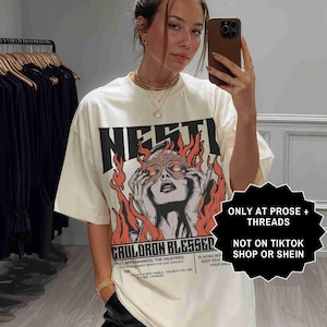 May include: Cream-colored graphic t-shirt with the word "NESTI" in bold black letters. The shirt features a graphic of a woman with flames and the text "CAULDRON BLESSED". The shirt also has the text "ONLY AT PROSE + THREADS" and "NOT ON TIKTOK SHOP OR SHEIN".