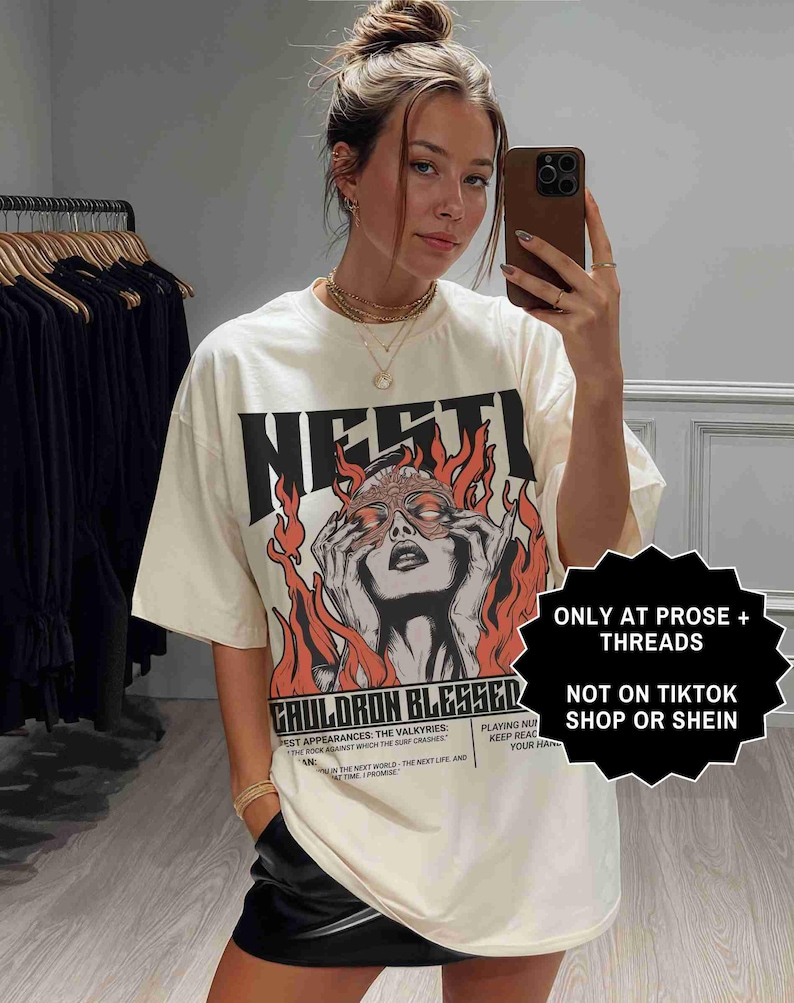 May include: Cream-colored graphic t-shirt with the word "NESTI" in bold black letters. The shirt features a graphic of a woman with flames and the text "CAULDRON BLESSED". The shirt also has the text "ONLY AT PROSE + THREADS" and "NOT ON TIKTOK SHOP OR SHEIN".