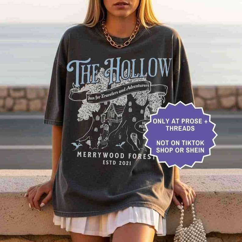Pu&ograve; includere: T-shirt grigio scuro con la scritta "The Hollow" in azzurro, con una grafica di albero e casa. La maglietta include anche il testo "Merrywood Forest Estd 2021".