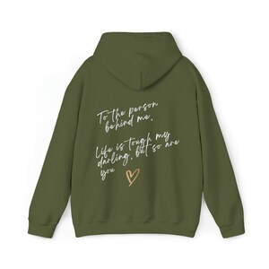 May include: Olive green hoodie with the text "To the person behind me, Life is tough my darling, but so are you" in a handwritten style, with a gold heart graphic. The hoodie has a hood and long sleeves.