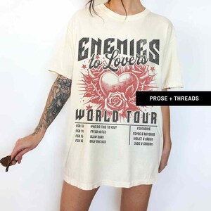 Pode incluir: Uma camiseta bege com uma estampa gr&aacute;fica preta e vermelha que diz "Enemies to Lovers World Tour". O gr&aacute;fico apresenta um cora&ccedil;&atilde;o com uma rosa e espinhos. A camiseta tamb&eacute;m lista as datas da turn&ecirc; e os artistas.