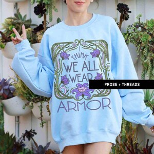 May include: Light blue sweatshirt with a floral design and the text "I think we all wear armor".