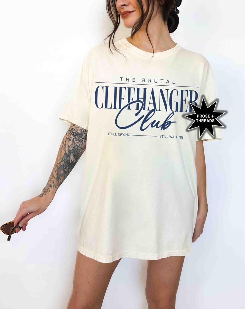 May include: A white t-shirt with the text "The Brutal Cliffhanger Club" in navy blue lettering. The text "Still Crying" and "Still Waiting" are printed below the main text. A black star with the text "Prose + Threads" is in the top right corner of the shirt.