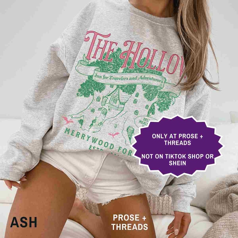 May include: A light grey sweatshirt featuring the text "The Hollow" in pink and green. The design includes a tree with a building and the words "Inn for Travelers and Adventurers". The sweatshirt also has the words "Merrywood For" and "Ash".