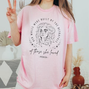 May include: A light pink t-shirt featuring a black and white illustration of Medusa with the text "HER WALLS WERE BUILT BY THE BETRAYALS of those she loved -MEDUSA-". The design is encircled by stars and a circular border.
