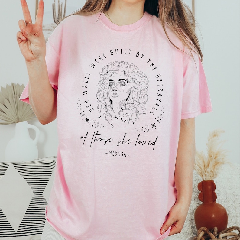 May include: A light pink t-shirt featuring a black and white illustration of Medusa with the text "HER WALLS WERE BUILT BY THE BETRAYALS of those she loved -MEDUSA-". The design is encircled by stars and a circular border.