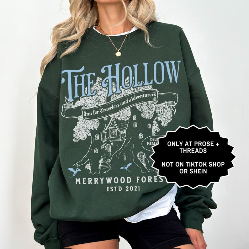 May include: Dark green sweatshirt featuring the text "THE HOLLOW" in light blue, with a banner reading "Inn for Travelers and Adventurers". Below is a treehouse design and the words "MERRYWOOD FOREST ESTD 2021".
