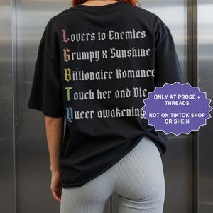 Pu&ograve; includere: Una t-shirt nera con testo bianco in carattere gotico sul retro, che elenca tropi letterari: Lovers to Enemies, Grumpy x Sunshine, Billionaire Romance, Touch her and Die e Queer awakening, con le lettere iniziali in un gradiente arcobaleno.
