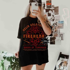 Pode incluir: Camiseta preta com um design gr&aacute;fico vermelho e laranja que diz "RELIN GALATHY FIREHEART QUEEN OF TERRASEN". A camisa tamb&eacute;m tem o texto "BATTLE THE STARS". Um bon&eacute; de beisebol preto &eacute; usado.