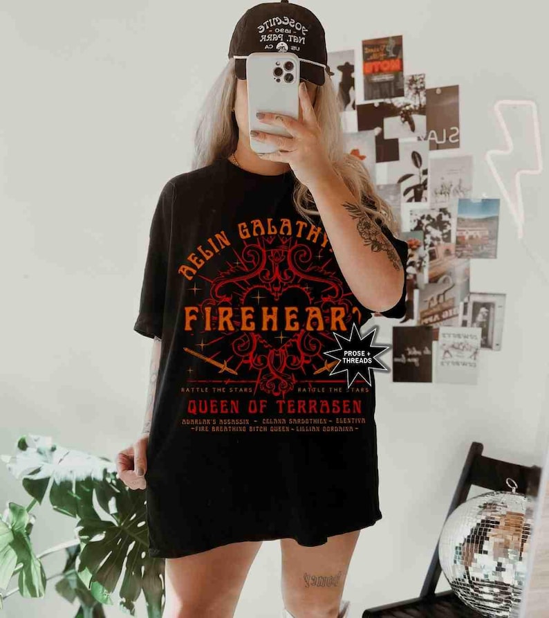 Pode incluir: Camiseta preta com um design gr&aacute;fico vermelho e laranja que diz "RELIN GALATHY FIREHEART QUEEN OF TERRASEN". A camisa tamb&eacute;m tem o texto "BATTLE THE STARS". Um bon&eacute; de beisebol preto &eacute; usado.