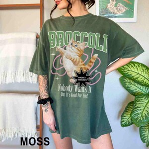 May include: Green oversized t-shirt with a graphic of a cat holding a pink string and the text "Broccoli" in white. The text "Prose + Threads" is in a black star shape. The text "Nobody Wants It But It's Good For You!" is below the cat graphic. The text "Moss" is in white on a brown surface.