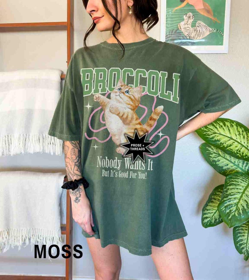 May include: Green oversized t-shirt with a graphic of a cat holding a pink string and the text "Broccoli" in white. The text "Prose + Threads" is in a black star shape. The text "Nobody Wants It But It's Good For You!" is below the cat graphic. The text "Moss" is in white on a brown surface.