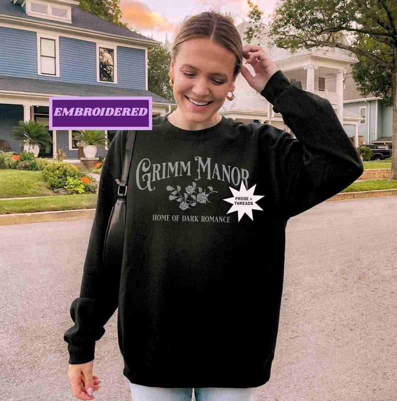 May include: Black crew neck sweatshirt featuring the text "Grimm Manor: Home of Dark Romance" in grey. The design includes floral and star graphics. The word "Embroidered" is in a purple rectangle.