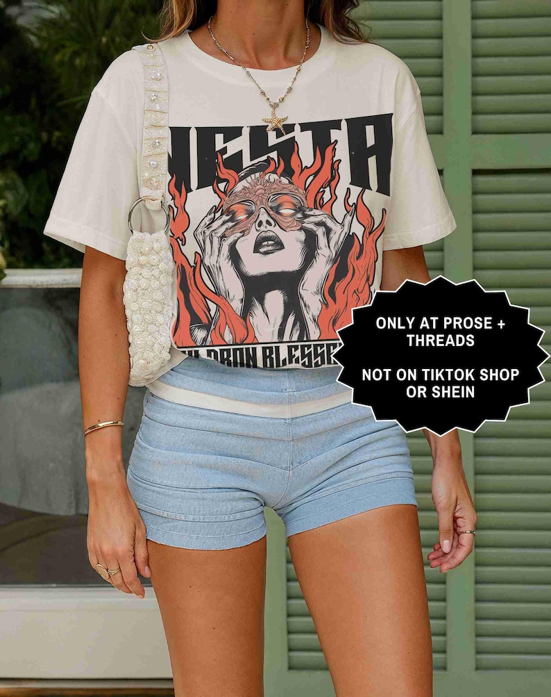 May include: Cream-colored graphic tee with a red and black illustration of a woman with flames. The tee is paired with light blue shorts. A white beaded purse and gold jewelry complete the look. Text on the tee reads "NEITH" and "BORN BLESSK".