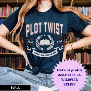 May include: A navy blue t-shirt with the text "PLOT TWIST" in white with red outlines. The shirt also features a white book with a laurel wreath and the text "Book Club" and "Expect the Unexpected". The shirt is being worn by a person with long brown hair. The text "100% of profits donated to LA WILDFIRE RELIEF" is in a pink starburst.