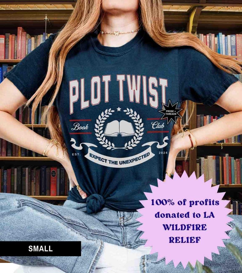 May include: A navy blue t-shirt with the text "PLOT TWIST" in white with red outlines. The shirt also features a white book with a laurel wreath and the text "Book Club" and "Expect the Unexpected". The shirt is being worn by a person with long brown hair. The text "100% of profits donated to LA WILDFIRE RELIEF" is in a pink starburst.