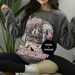 May include: A gray sweatshirt with a graphic print of a haunted house, ghosts, and a cat. The text on the sweatshirt reads "Salem Haunted Library Massachusetts" and "Prose + Threads".