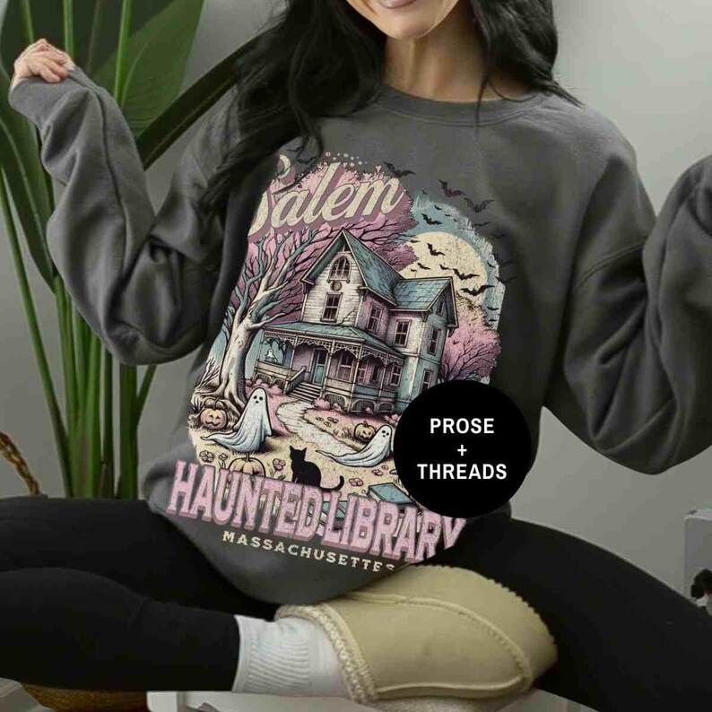 May include: A gray sweatshirt with a graphic print of a haunted house, ghosts, and a cat. The text on the sweatshirt reads "Salem Haunted Library Massachusetts" and "Prose + Threads".