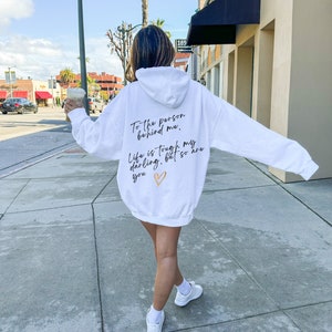 May include: White hoodie with the text "To the person behind me, Life is tough my darling, but so are you" in black script and a gold heart. The hoodie is worn with white sneakers.