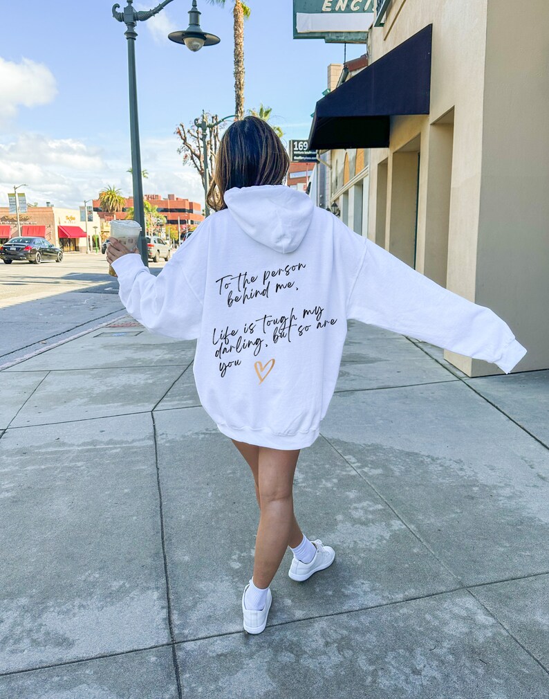May include: White hoodie with the text "To the person behind me, Life is tough my darling, but so are you" in black script and a gold heart. The hoodie is worn with white sneakers.