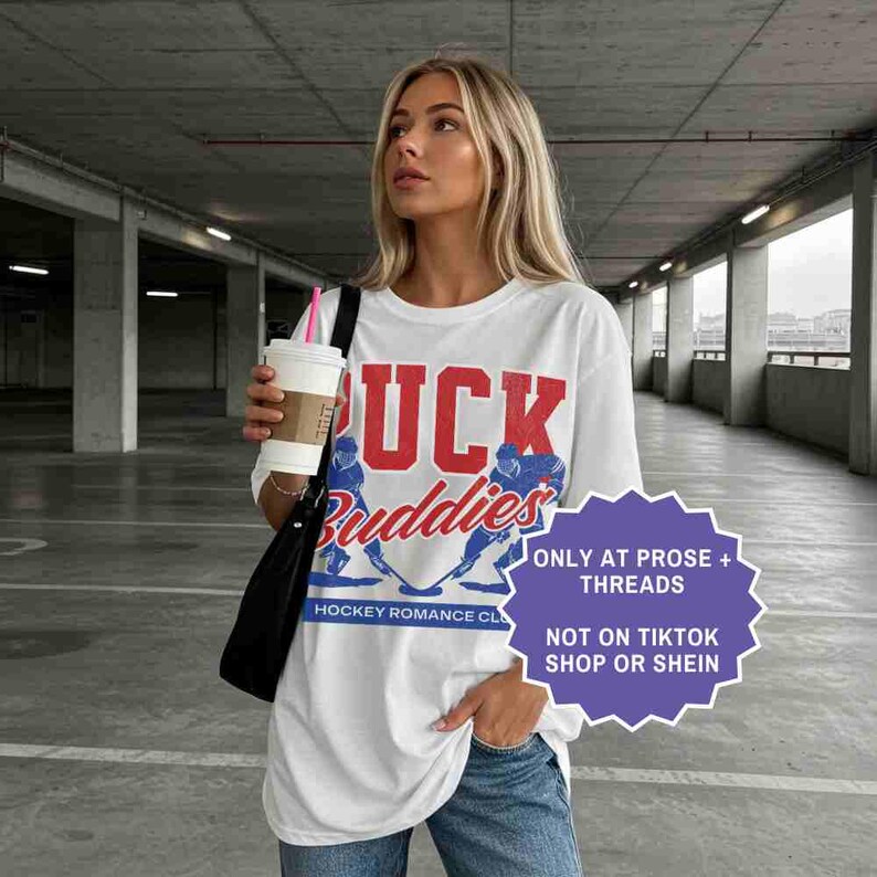 May include: White t-shirt with the text "PUCK Buddies" in red and blue, featuring hockey players. The shirt also has the text "HOCKEY ROMANCE CLUB" and a purple starburst with text.