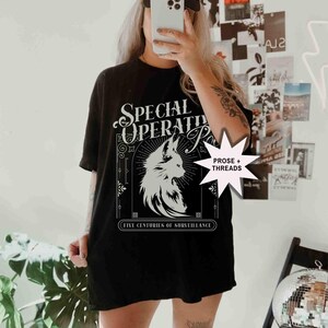May include: Black t-shirt with a white graphic of a fox and the words "SPECIAL OPERATIONS" and "FIVE CENTURIES OF SURVEILLANCE". The shirt is paired with a black baseball cap.