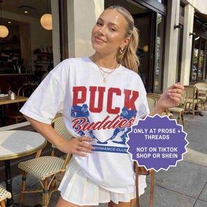 May include: White t-shirt with the text "PUCK Buddies" in red and blue, featuring hockey player graphics. The shirt is paired with a white pleated skirt. A purple sticker reads "ONLY AT PROSE + THREADS NOT ON TIKTOK SHOP OR SHEIN."