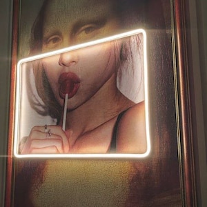 May include: A framed painting of a woman with brown hair, wearing a black top, and holding a lollipop in her mouth. The painting is lit with a white neon light.