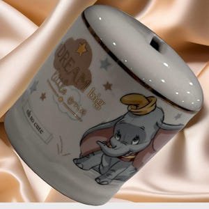 Puede incluir: Huchita de cerámica blanca con ribete dorado y una imagen de dibujos animados de Dumbo el elefante. La huchita tiene las palabras "Dream big little one" y "oh so cute" escritas en ella.