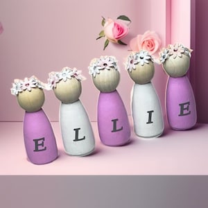May include: Five wooden peg dolls with floral crowns and the name "ELLIE" spelled out on their fronts. The dolls are painted in shades of purple and white.