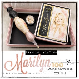 Op de afbeelding: Een herdenkingsset met een zwarte en roségouden pen met de tekst Marilyn 1926-2026, een bijpassend rechthoekig blik met een portret van Marilyn Monroe en een kleine ovale speld, alles gepresenteerd in een lichtroze dienblad met zwarte voering.