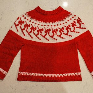 Puede incluir: Un jersey rojo tejido con un patr&oacute;n blanco alrededor del canes&uacute; y los pu&ntilde;os. El patr&oacute;n blanco presenta figuras estilizadas. El jersey tiene cuello redondo y mangas largas. El dobladillo inferior y los pu&ntilde;os tienen un borde decorativo blanco.