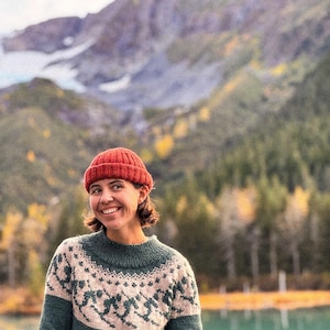 Puede incluir: Una mujer con un gorro de punto rojo y un su&eacute;ter verde y beige con estampado sonr&iacute;e frente a un lago y un paisaje monta&ntilde;oso. El su&eacute;ter presenta un dise&ntilde;o fair isle alrededor del canes&uacute; y las mangas.