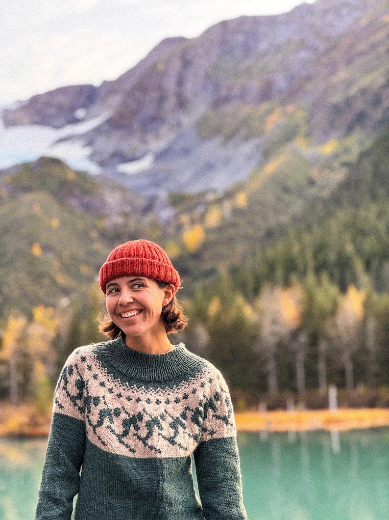 Puede incluir: Una mujer con un gorro de punto rojo y un su&eacute;ter verde y beige con estampado sonr&iacute;e frente a un lago y un paisaje monta&ntilde;oso. El su&eacute;ter presenta un dise&ntilde;o fair isle alrededor del canes&uacute; y las mangas.
