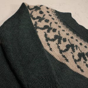 Puede incluir: Jersey de punto verde oscuro con un dise&ntilde;o beige estampado en la parte superior del pecho y los hombros. El jersey tiene mangas largas y un pu&ntilde;o acanalado. El patr&oacute;n de punto es visible en primer plano.