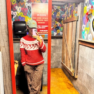 Puede incluir: Un su&eacute;ter rojo y blanco estampado se refleja en un espejo con marco rojo. El espejo est&aacute; en una habitaci&oacute;n con arte de graffiti y una puerta de madera. La persona en el reflejo lleva un gorro verde y pantalones marrones estampados.
