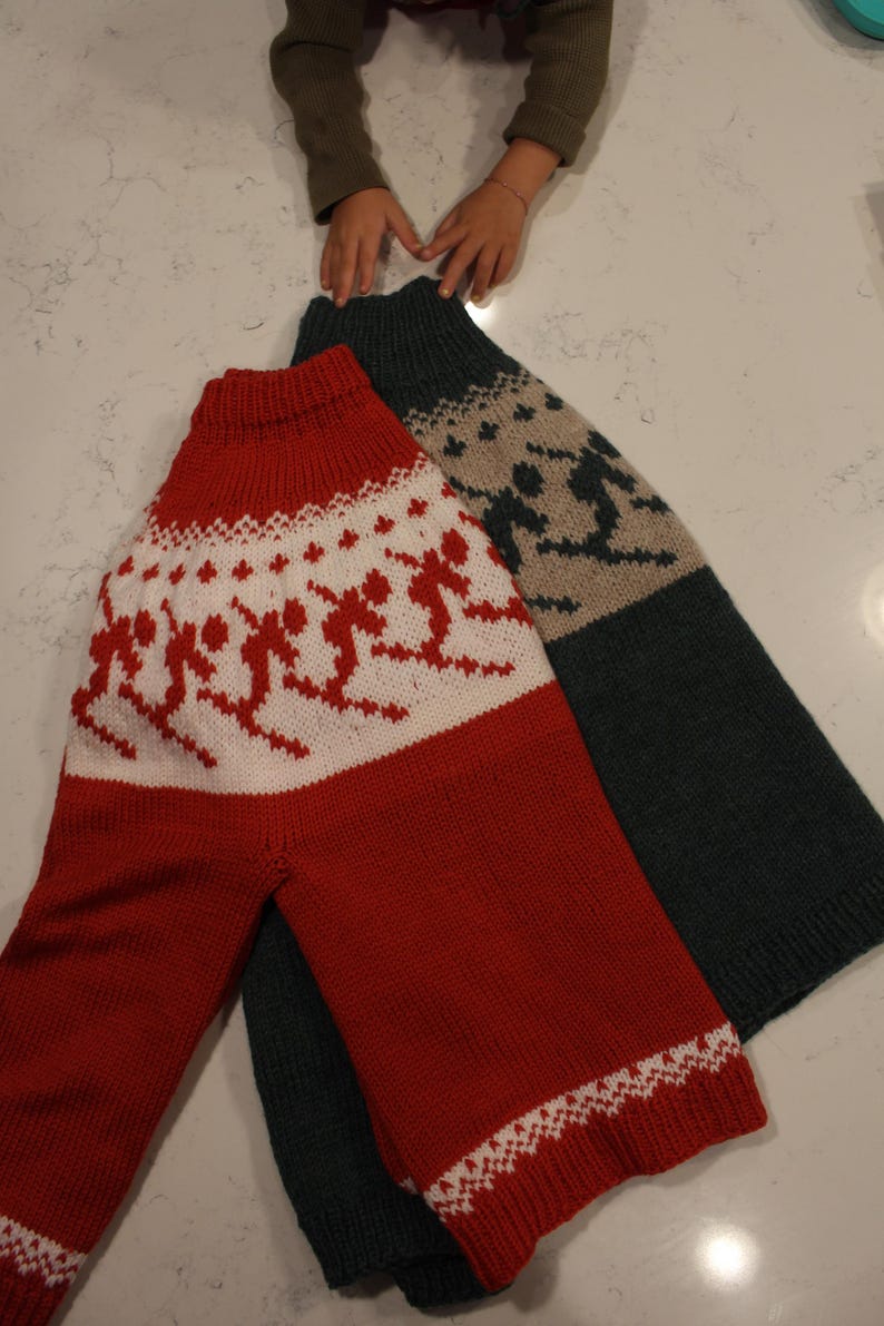 Puede incluir: Dos su&eacute;teres de punto. Uno es rojo con esquiadores blancos y un borde blanco estampado. El otro es verde oscuro con un estampado de esquiador beige. Ambos su&eacute;teres tienen cuello alto y mangas largas.